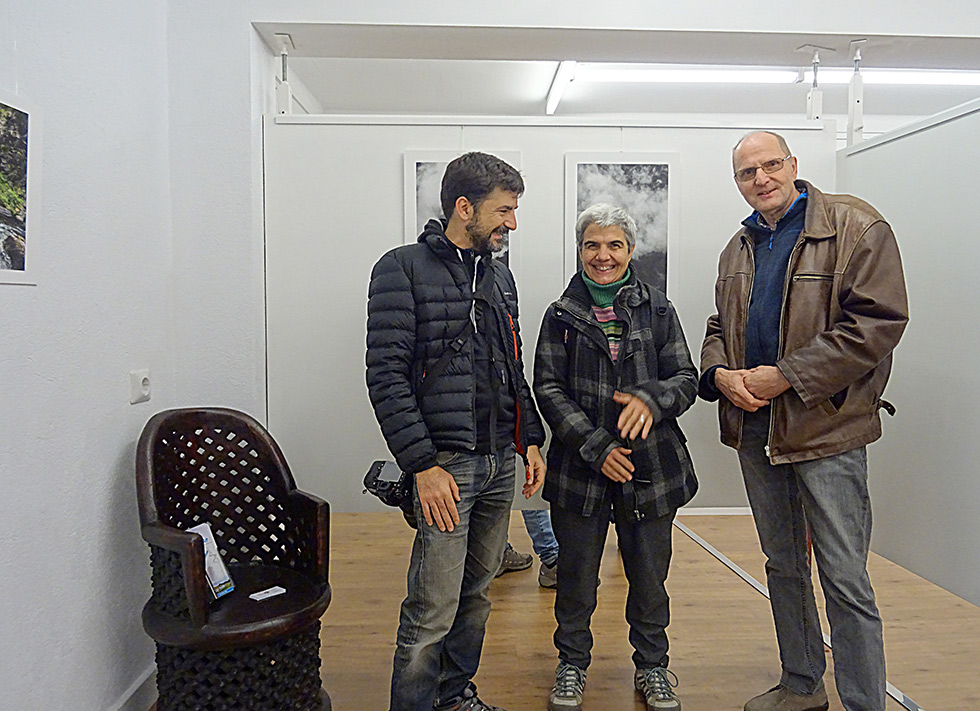 Three people smiling and conversing in a gallery with framed photographs on the wall.