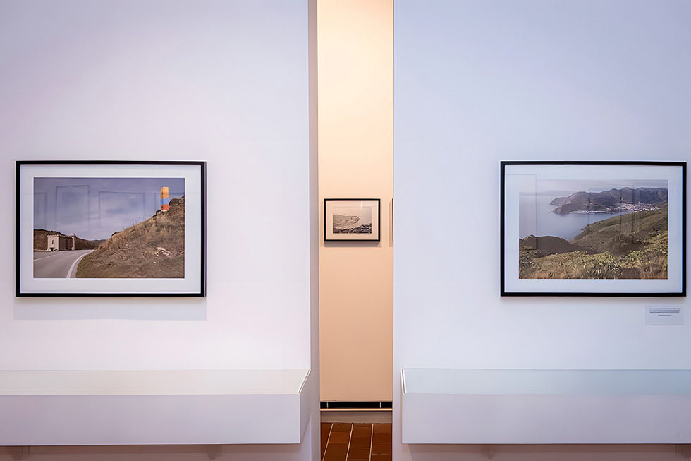 Two framed landscape photographs are displayed on a white wall with a small black and white photograph visible in the background through an opening.