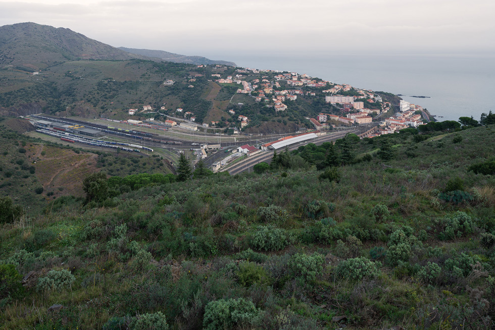 A view of a coastal town with a large railway station with multiple stub tracks running through a valley and houses scattered on the hillside, extending towards the sea.