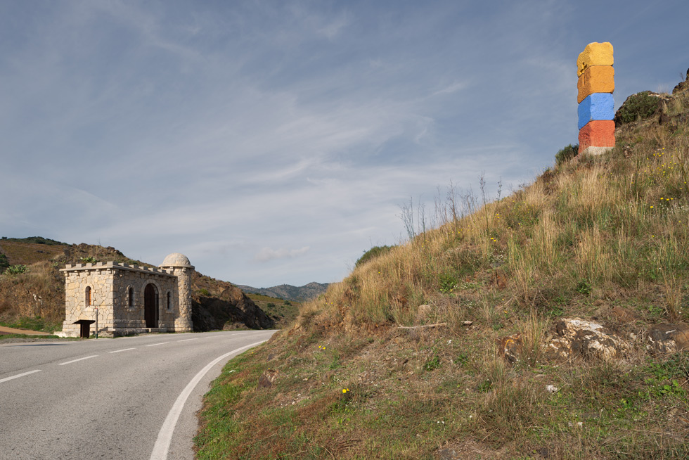 A winding road with a minuscule stone building resembling a castle on the left and a colorful stacked pillar on the right, set against a backdrop of rolling hills under a blue sky.