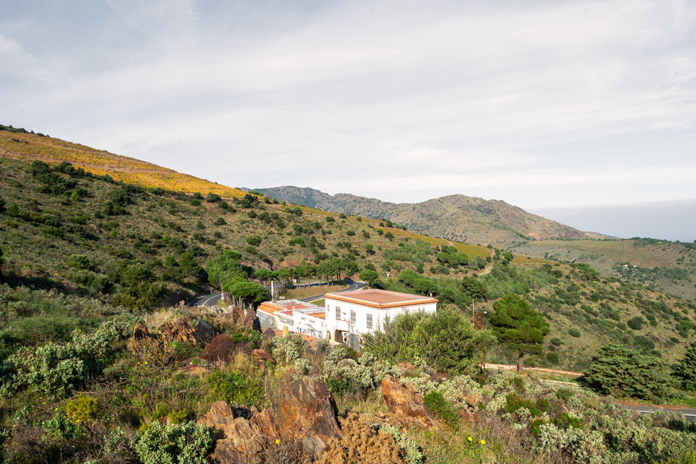 A scenic view of a white building surrounded by hills covered in bushes and different cactuses, with a winding road and a mountainous backdrop under a partly cloudy sky.