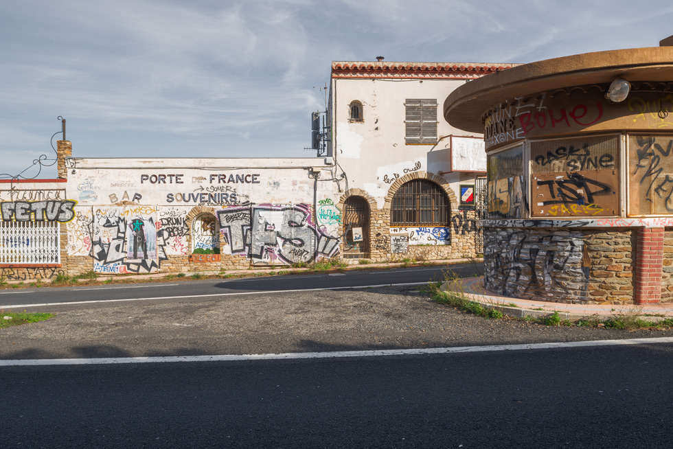 An abandoned building with 'Porte de France, Souvenirs' written on the wall, covered in graffiti in front an empty road.