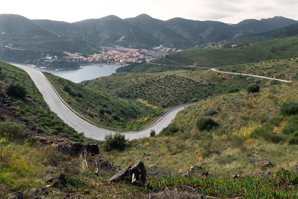 A winding road leading through green hills, covered in bushes and rocks, towards a coastal town nestled between mountains and the sea.