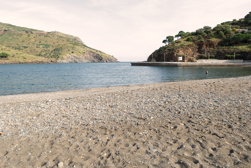 A quiet pebble beach with tranquil blue waters, surrounded by green hills and rocky outcrops.