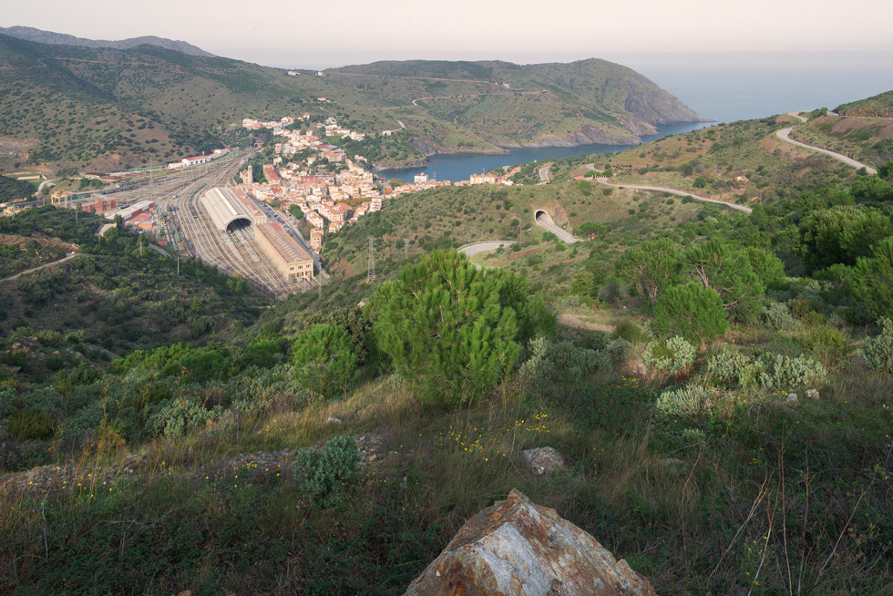 A scenic view of a coastal town with a huge railway station with multiple stub tracks, surrounded by green hills and overlooking a serene bay.