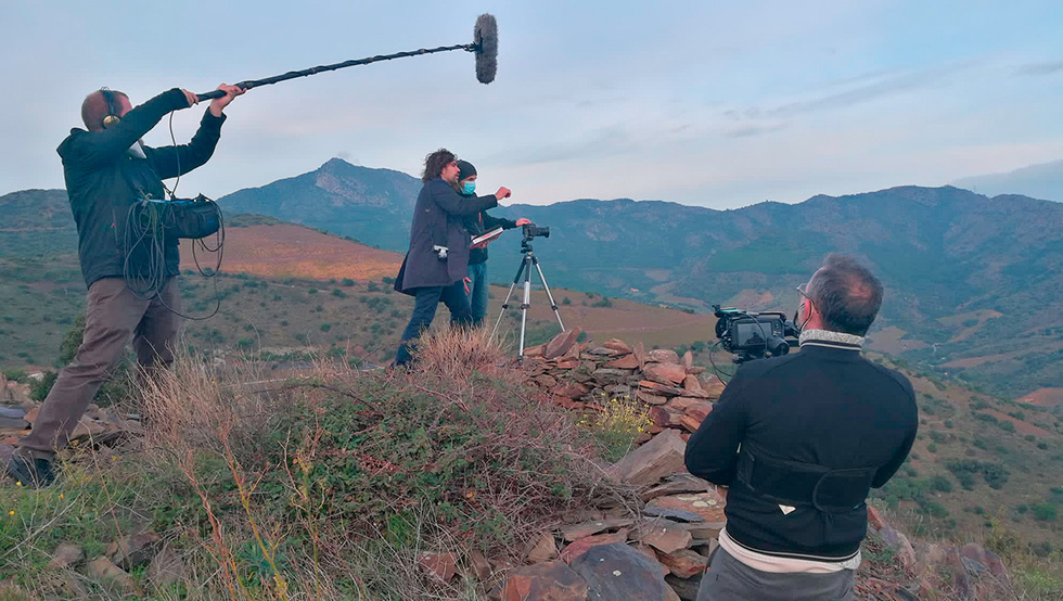 A film crew shooting on a hillside with a scenic mountain backdrop. The crew includes a cameraman and a person holding a boom microphone, they are filming two people setting up a tripod with a camera.