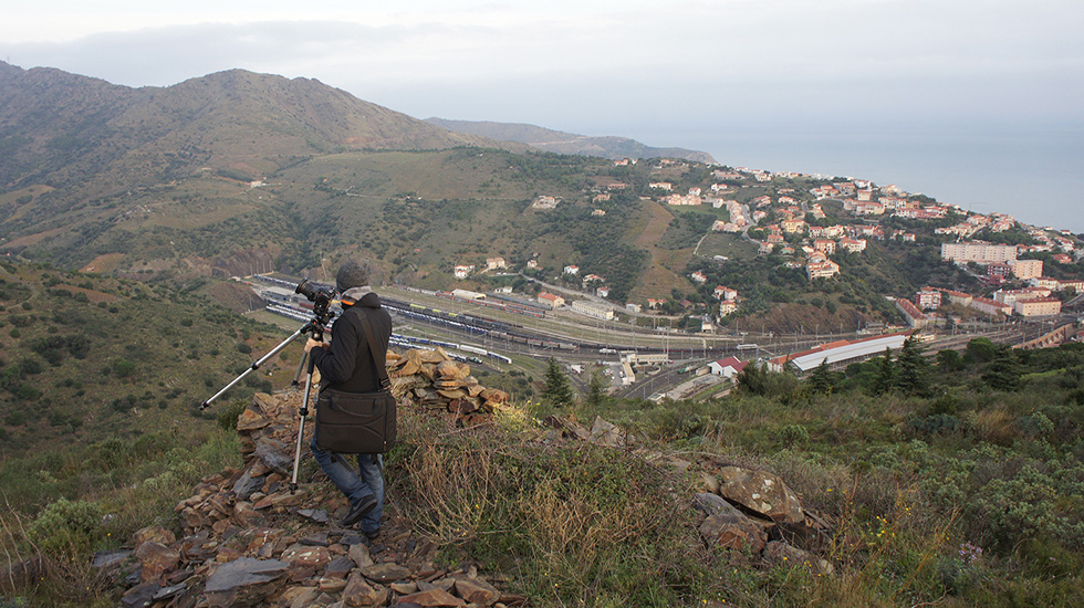 A photographer with a tripod about to capture a scenic view of a coastal town with a large railway station nestled among hills.