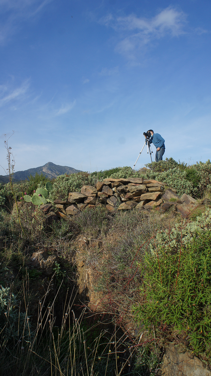 A photographer using a tripod on a rocky hillside, capturing a landscape scene with mountains in the background under a clear blue sky.