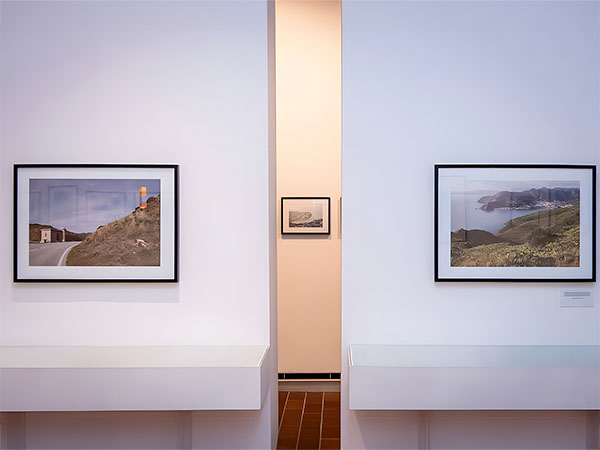 Two framed landscape photographs are displayed on a white wall with a small black and white photograph visible in the background through an opening.