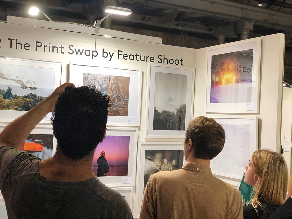 Three people viewing framed photographs on display at 'The Print Swap by Feature Shoot' exhibition.