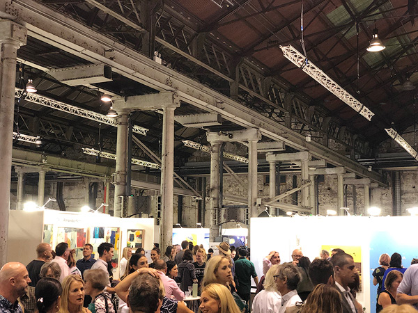 A bustling art exhibition with many people viewing various artworks in a large industrial-style space with high ceilings and exposed beams.