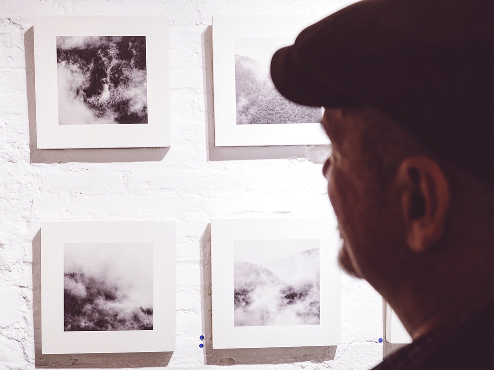 A person looking at a series of black and white square photographs of clouds and mist on a white brick wall.