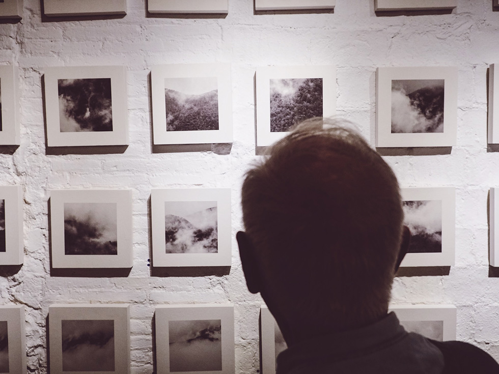 A person viewing a large collection of black and white square photographs of clouds and mist on a white brick wall.