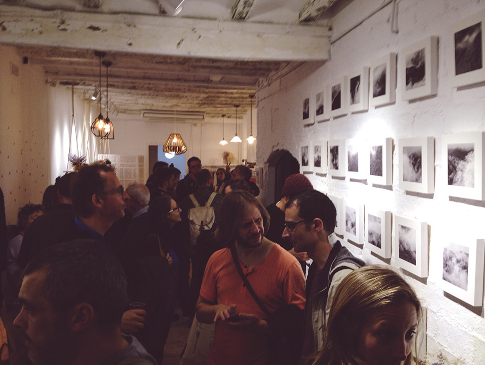 A crowded art gallery with people viewing black and white photographs of clouds and mist arranged in a grid on a white brick wall.