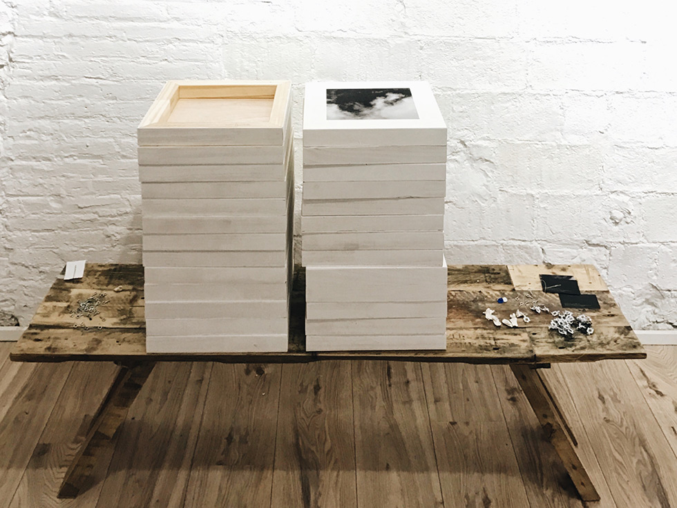 Two stacks of white-framed photographs on a wooden table, next to small metal hooks and other hanging materials, in a gallery with white brick walls.