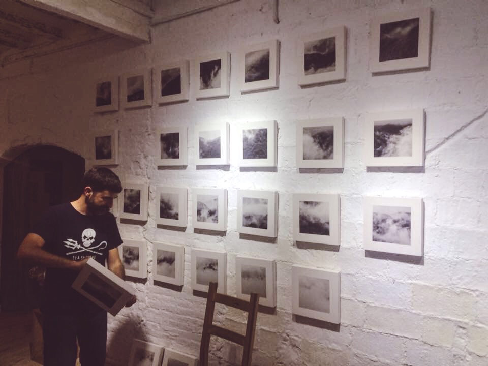 A man arranging black-and-white photographs on a white brick wall, with several already displayed in a grid pattern, in a gallery setting.