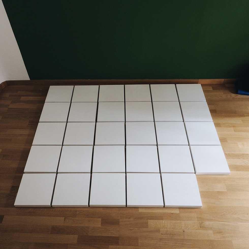 A grid of white square canvases laid out on a wooden floor.