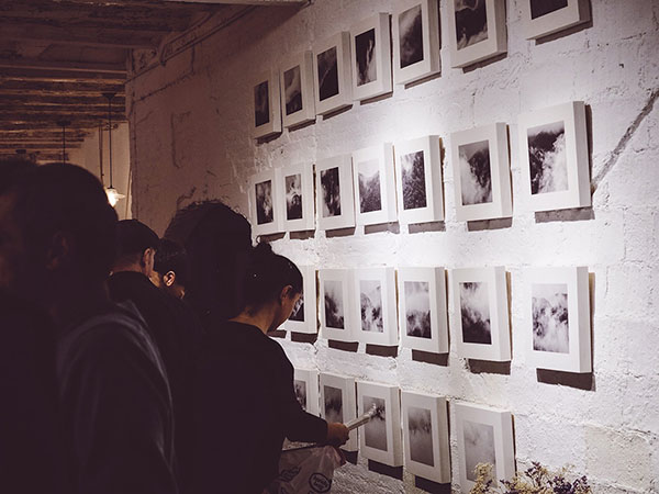 People viewing a series of black and white photographs of clouds and mist arranged in a grid on a white brick wall.