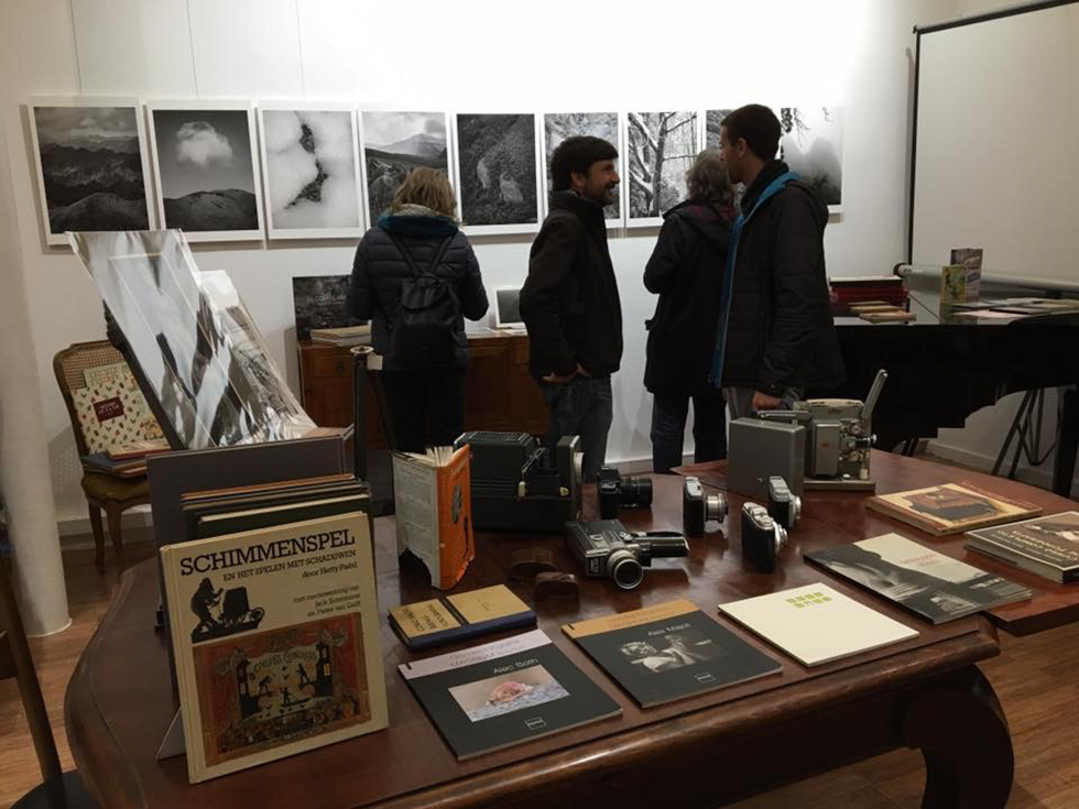 A group of people view black-and-white photographs on display in a cozy exhibition space. The room features a wooden table with vintage cameras, books, and other items. The photographs depict mountainous landscapes and other natural scenes.