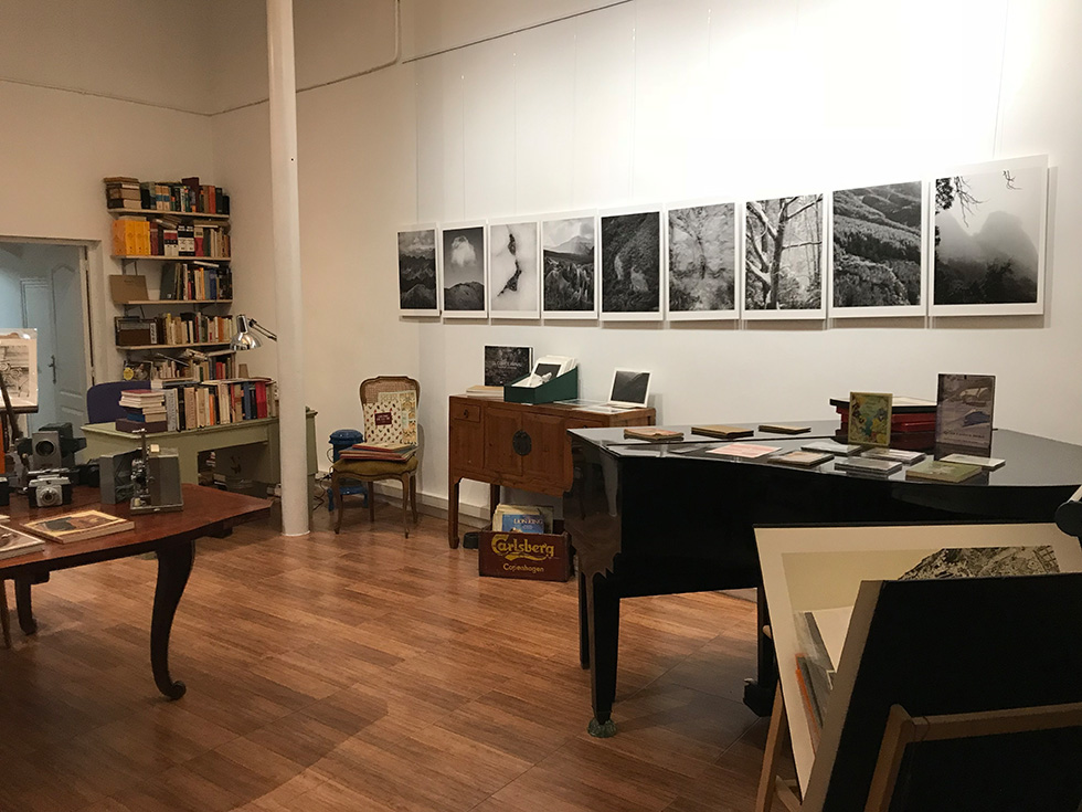 An exhibition space with black-and-white photographs of mountainous landscapes displayed on a wall, a piano with books and pamphlets on top, a desk with vintage cameras, and bookshelves in the background.