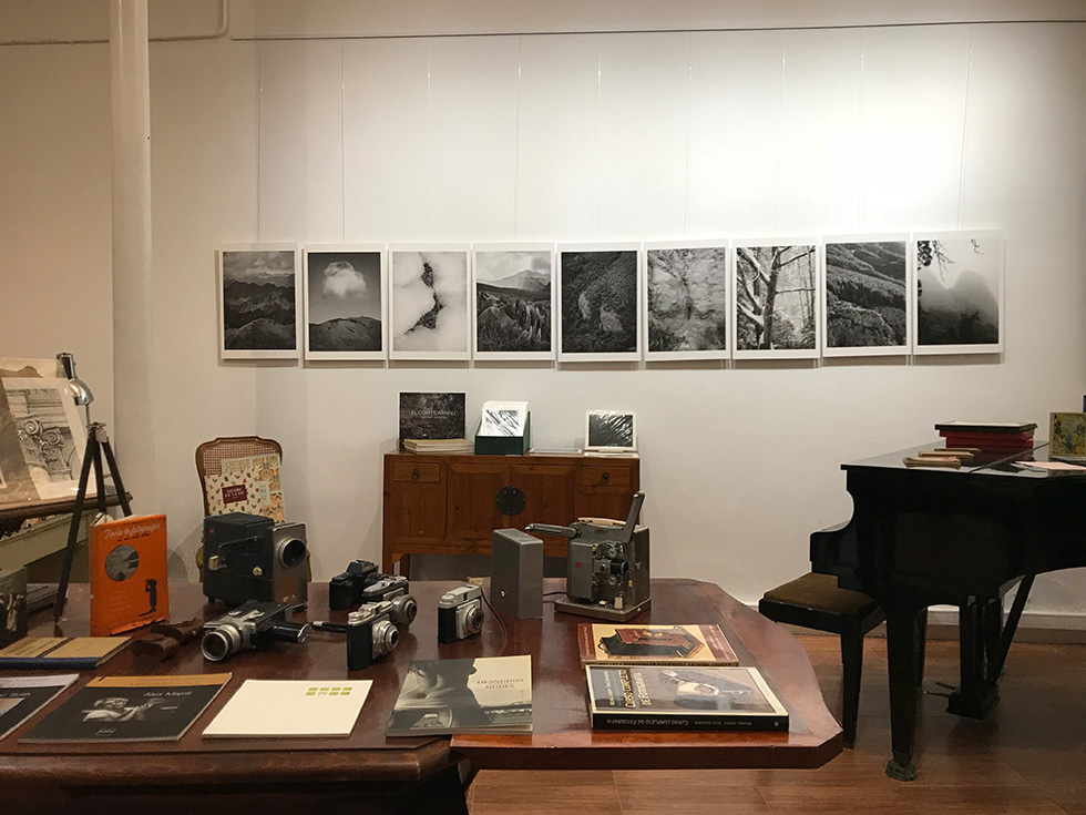 An exhibition space featuring black-and-white photographs of mountains on a wall, a table with vintage cameras and photography books, a piano, and various other books and items on display.