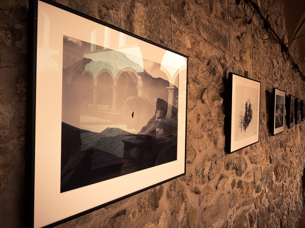 A close-up of framed photographs hanging on a stone wall in a gallery, showcasing various artworks in a row, the cloister's arches reflected on the photographs's glass.