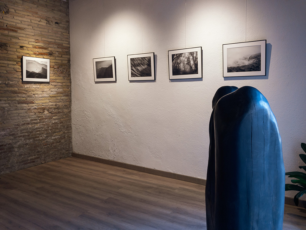 Photography exhibition with black and white landscape photos on white and brick walls, featuring a dark sculpture in the foreground.