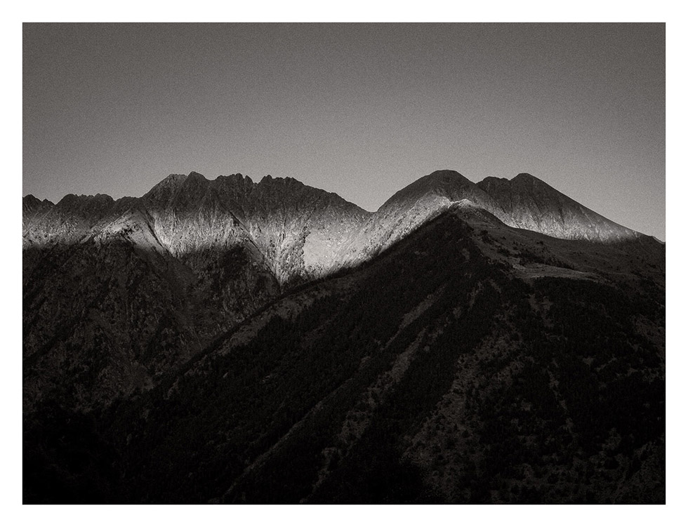 A mountain range just before sunset. Mountains on my back cast a deep shadow on them. Some clouds off scene partially block the sun, leaving the mountain ridge also in shadow. A thin strip of light illuminates the mountain range, right below the ridge.