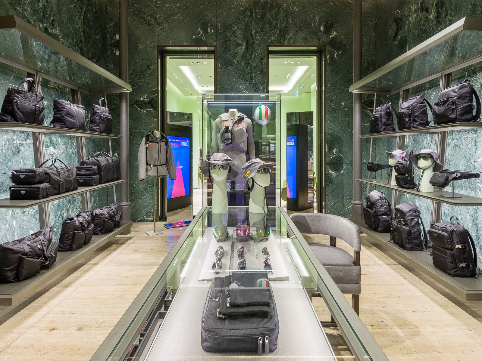 Lujosa boutique Prada con paredes de mármol verde, mostrando zapatillas de diseño, bolsos negros y accesorios en elegantes estanterías metálicas. Una mesa de vidrio exhibe gafas y sombreros, mientras que un maniquí en un atuendo de rendimiento está encerrado en una vitrina de vidrio.