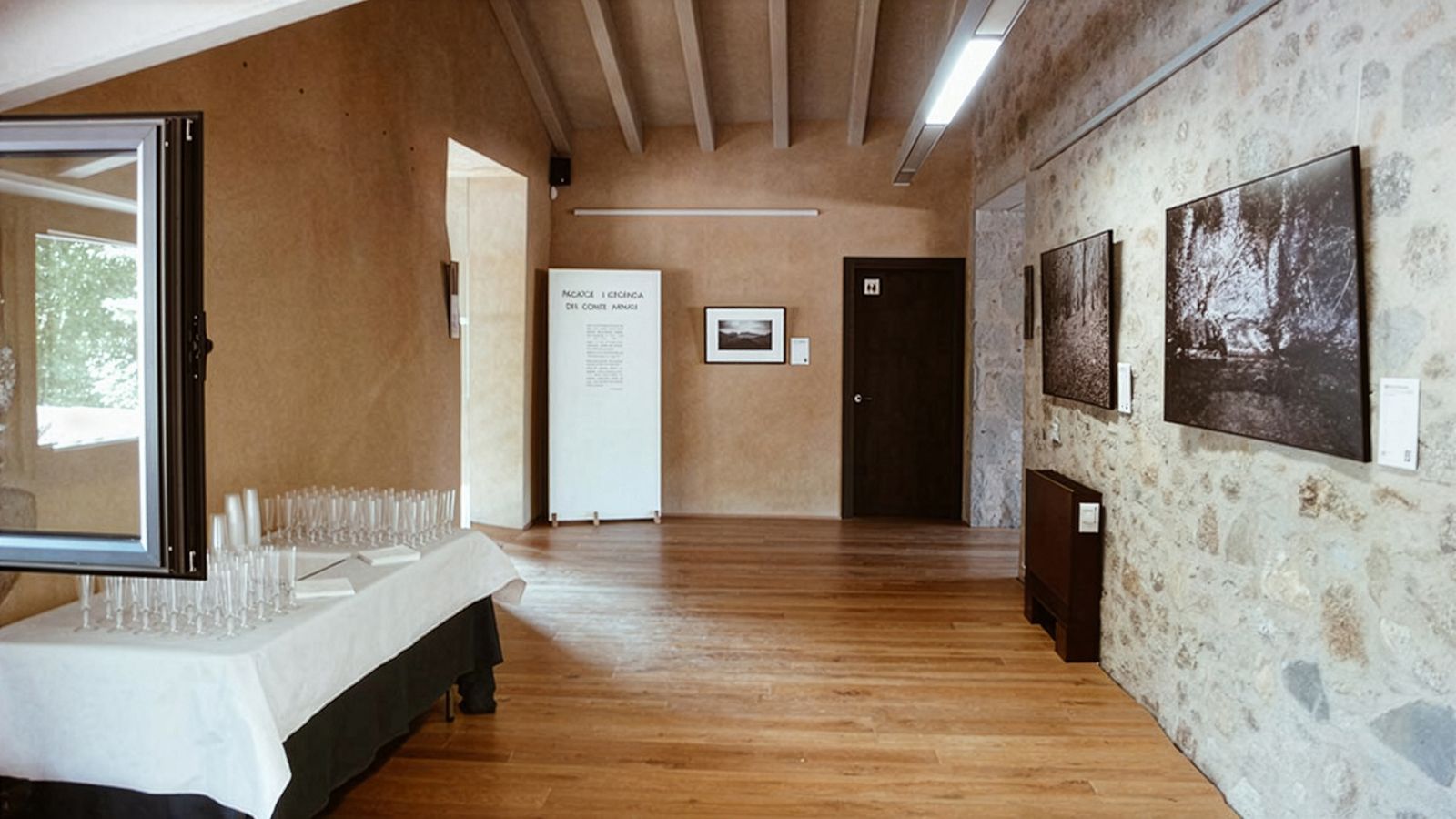 Sala con fotos enmarcadas en paredes de piedra y una mesa preparada con copas para el día de apertura.