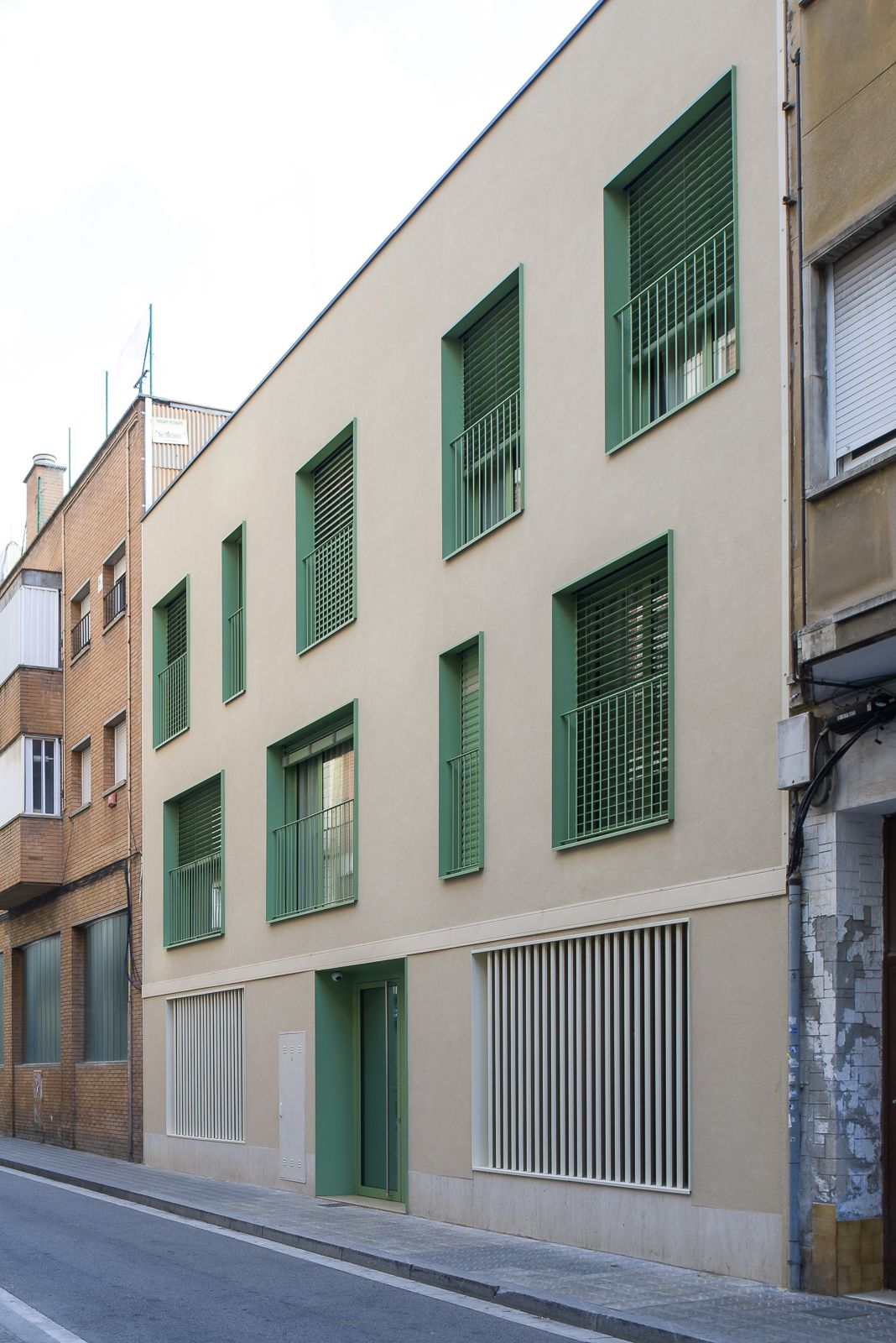 Low-angle view of a contemporary three-story building featuring light beige plaster walls, vibrant sage green window frames with integrated louvered blinds, and vertical white metal security bars over the ground floor windows.