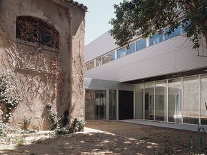 Un patio que combina arquitectura antigua y nueva, con un edificio histórico a la izquierda y una estructura moderna de vidrio a la derecha, rodeado de vegetación y árboles que dan sombra.