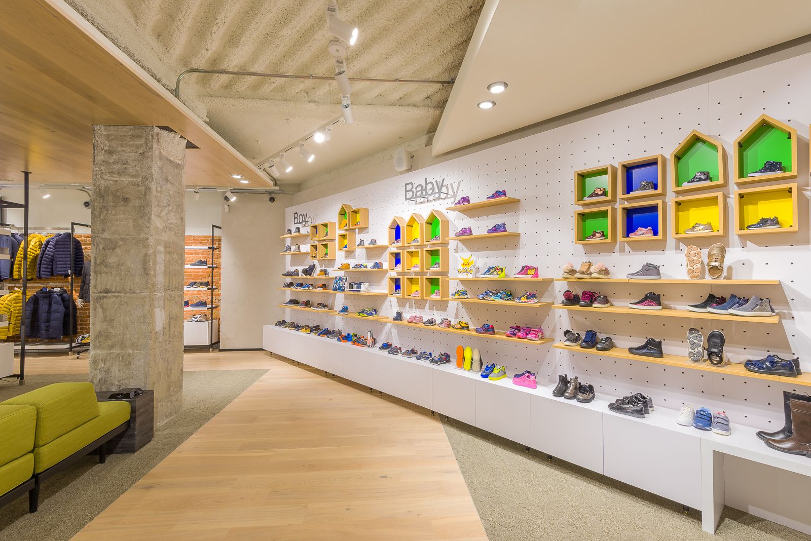 Sección de zapatos infantiles en la tienda Geox, con zapatos coloridos exhibidos en estantes y nichos de pared en forma de casa vibrantes, con un banco verde e iluminación moderna.