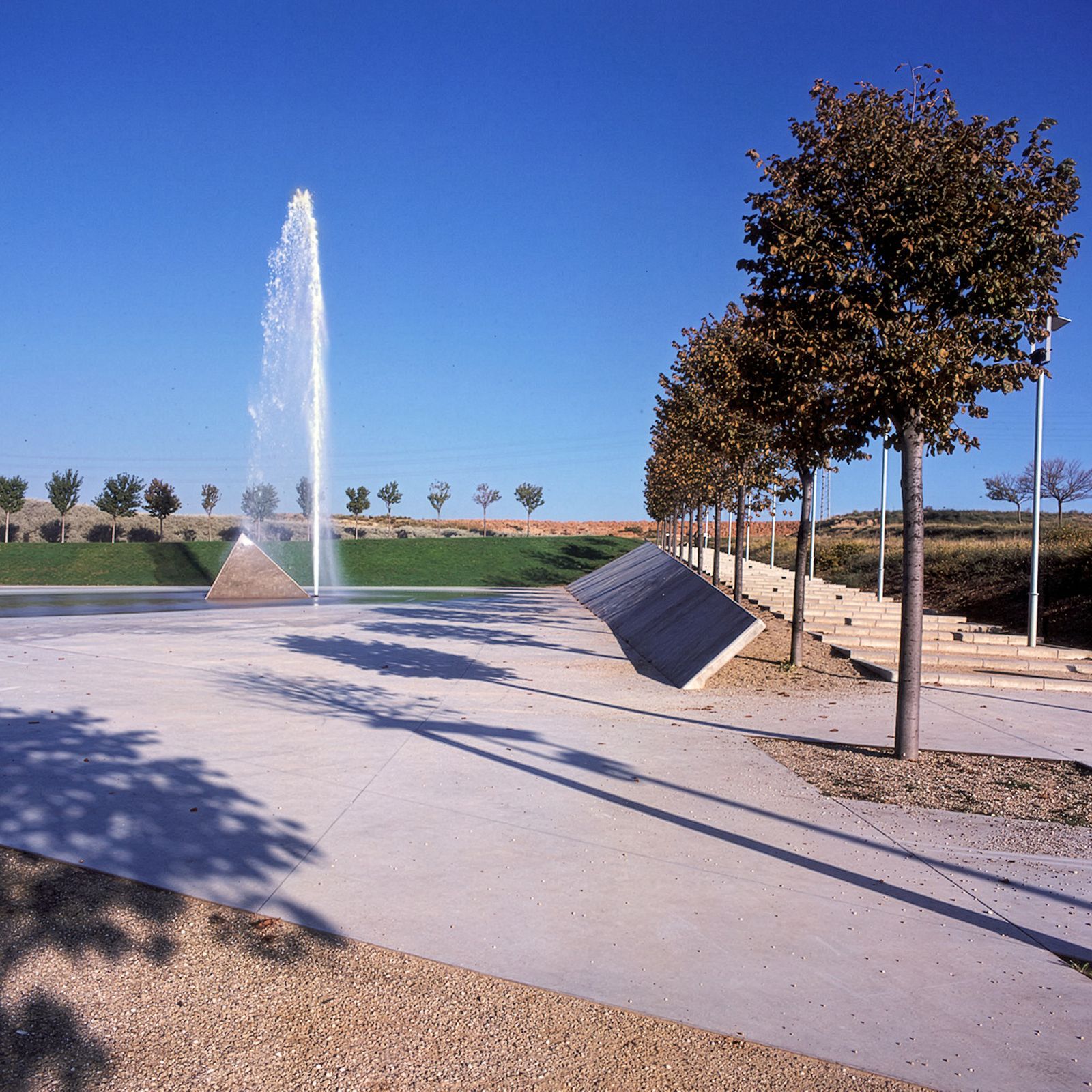 A line of trees along a walkway with gentle steps besides a large jet water fountain in an open park area.