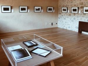Vista hacia una sala de galería con vigas de madera, fotos enmarcadas en las paredes y vitrinas sobre soportes de madera.