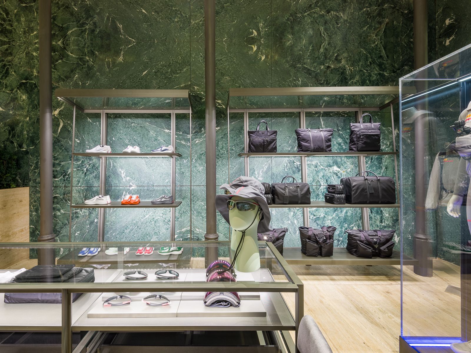 Boutique de lujo con paredes de mármol verde, mostrando zapatillas de diseño, bolsos negros y accesorios en elegantes estanterías metálicas. Una mesa de vidrio exhibe gafas y sombreros, mientras que un maniquí en un atuendo de rendimiento está encerrado en una vitrina de vidrio.