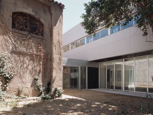 Un pati que combina arquitectura antiga i nova, amb un edifici històric a l'esquerra i una estructura de vidre moderna a la dreta, envoltats de vegetació i arbres que fan ombra.