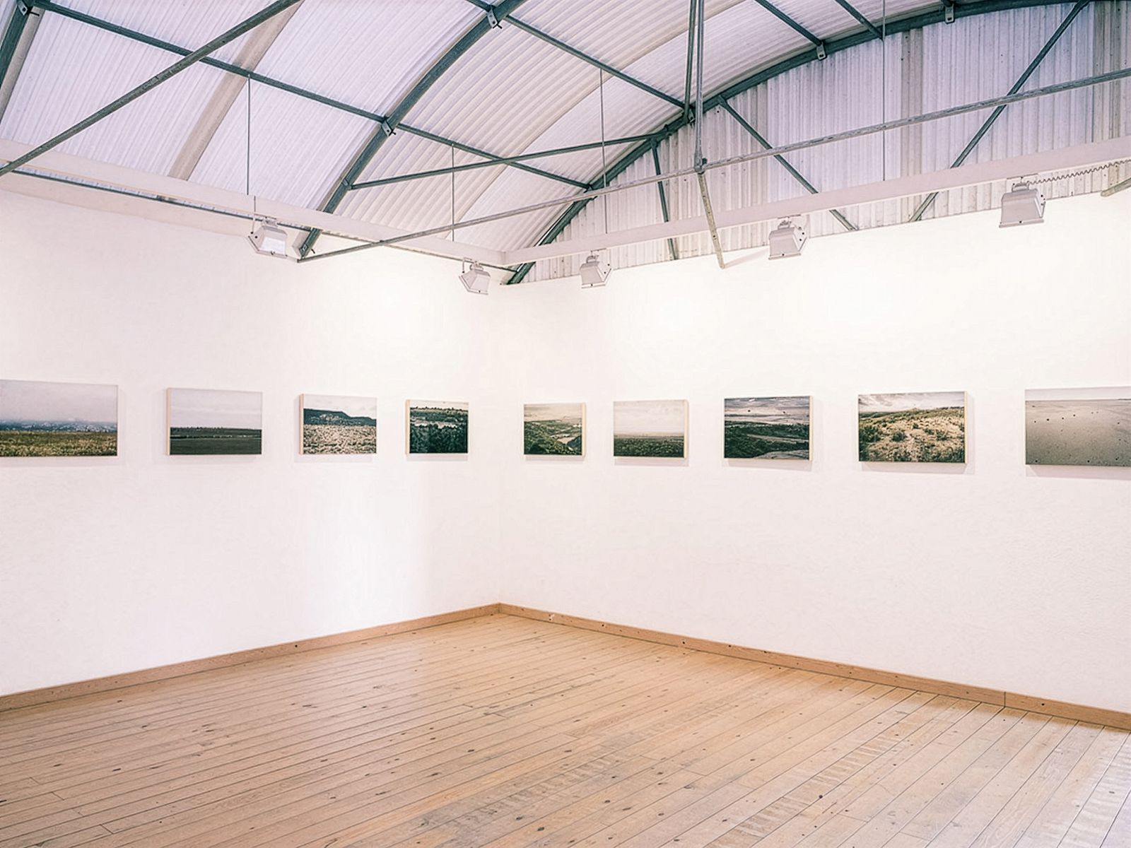 Exhibition hall with a series of landscape photographs arranged in a single row along the white walls, featuring a high curved ceiling and wooden floors.