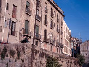 Edifici residencial antic amb una façana desgastada, amb balcons i situat sobre un talús de pedra. Al fons un edifici residencial nou seguint el patró orgànic del carrer