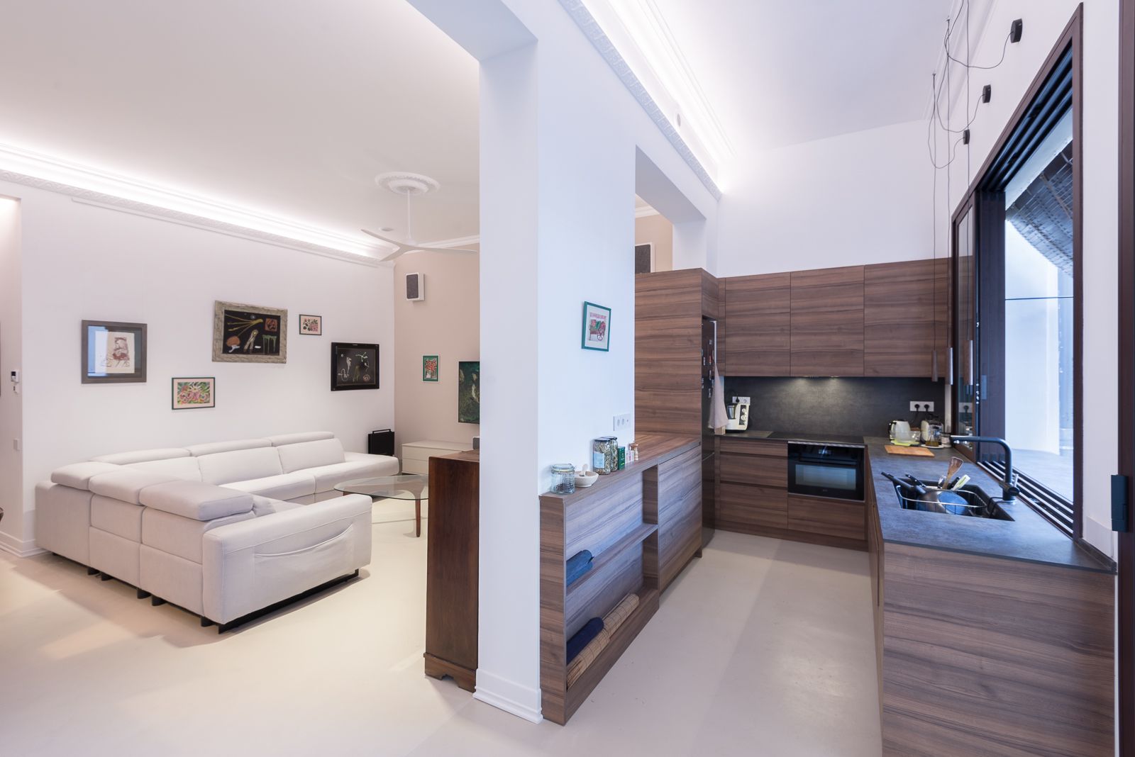 Modern open-concept living area with a cozy white sectional sofa, adjacent to a sleek wooden kitchen with built-in appliances.