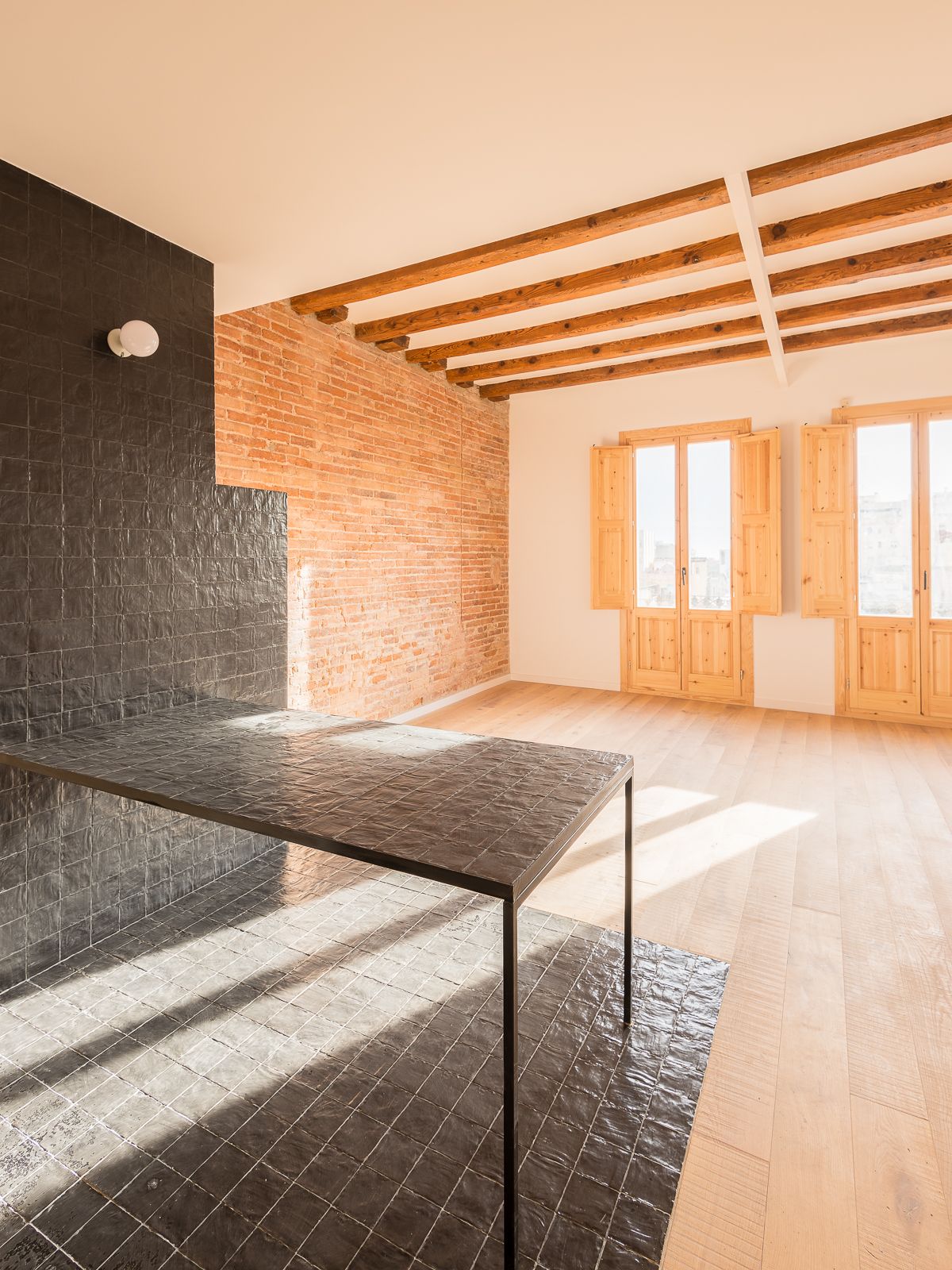 Área de estar de planta abierta con encimera de baldosas negras, pared de ladrillo expuesto, vigas de techo de madera y luz solar natural que entra a través de dos grandes balcones con persianas de madera.