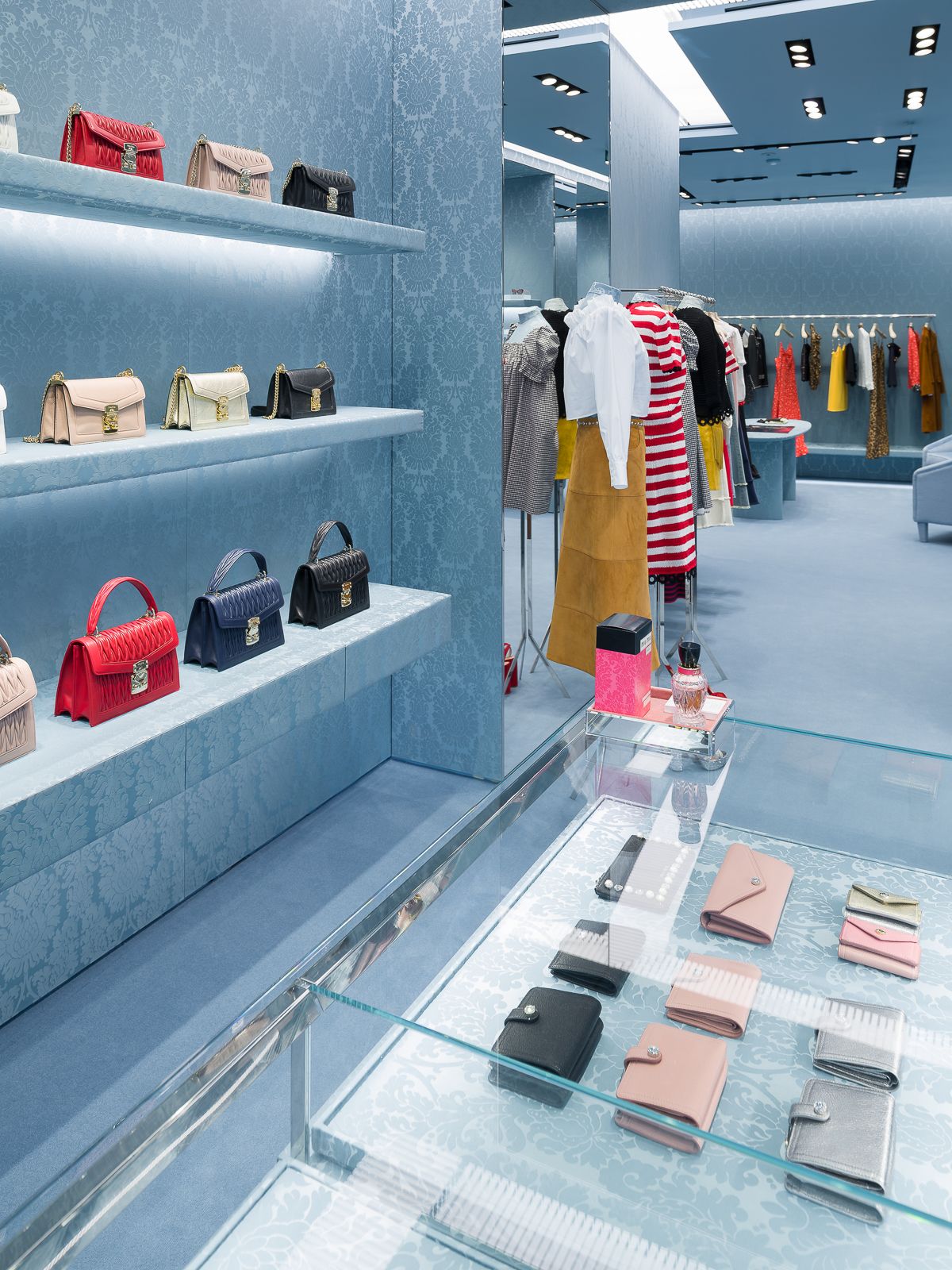 Interior de tienda Miu Miu con estantes con bolsos, un mostrador de exhibición de vidrio con carteras y percheros de ropa con varias prendas.