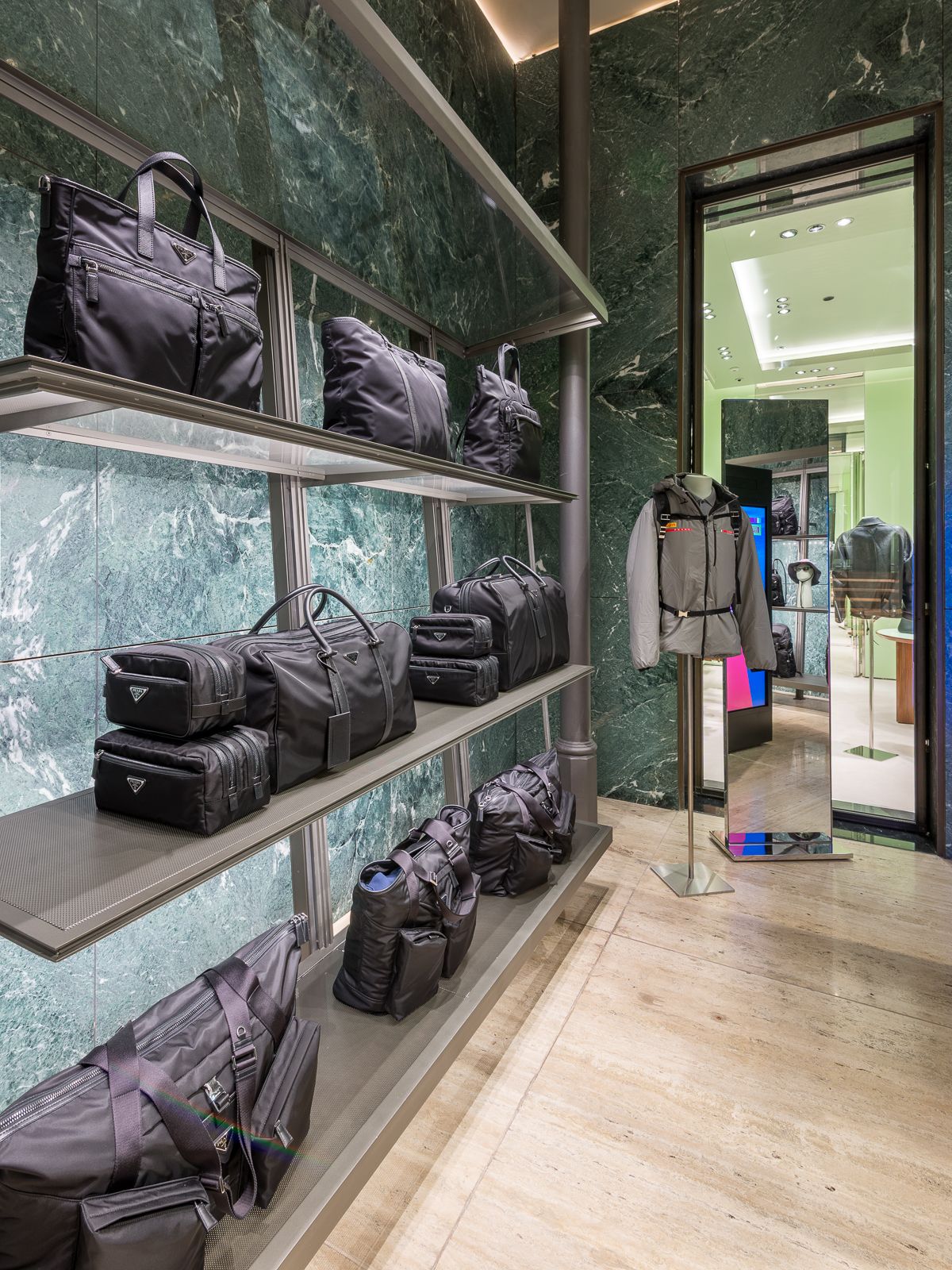 Lujosa boutique Prada con paredes de mármol verde, mostrando bolsos negros de diseño en elegantes estanterías metálicas. Un maniquí con prendas grises y una mochila junto a un espejo y una abertura a otra sección.