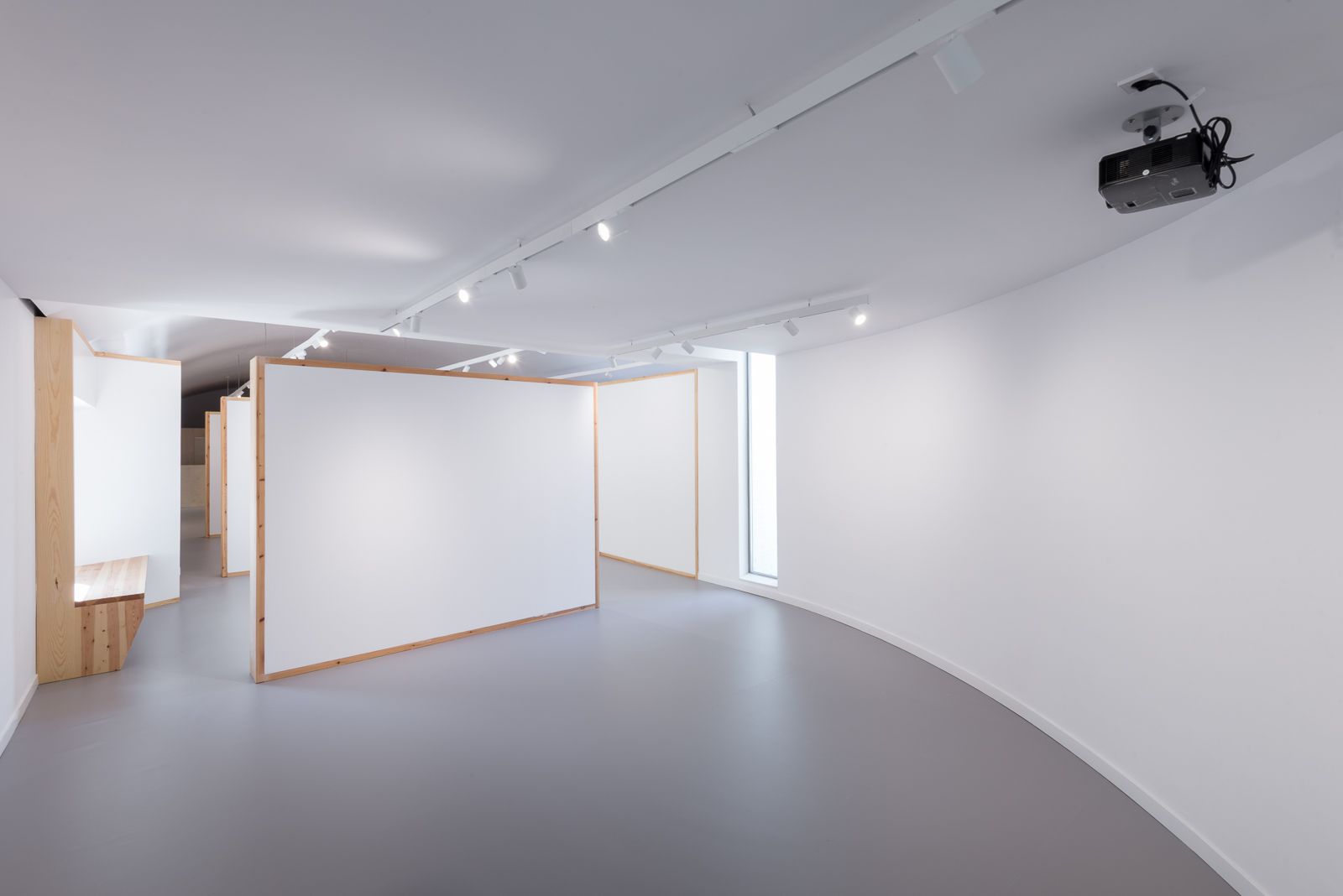Curved gallery space with modular white walls and wooden frames, a bench built into the wall, overhead track lighting, and a ceiling-mounted projector.
