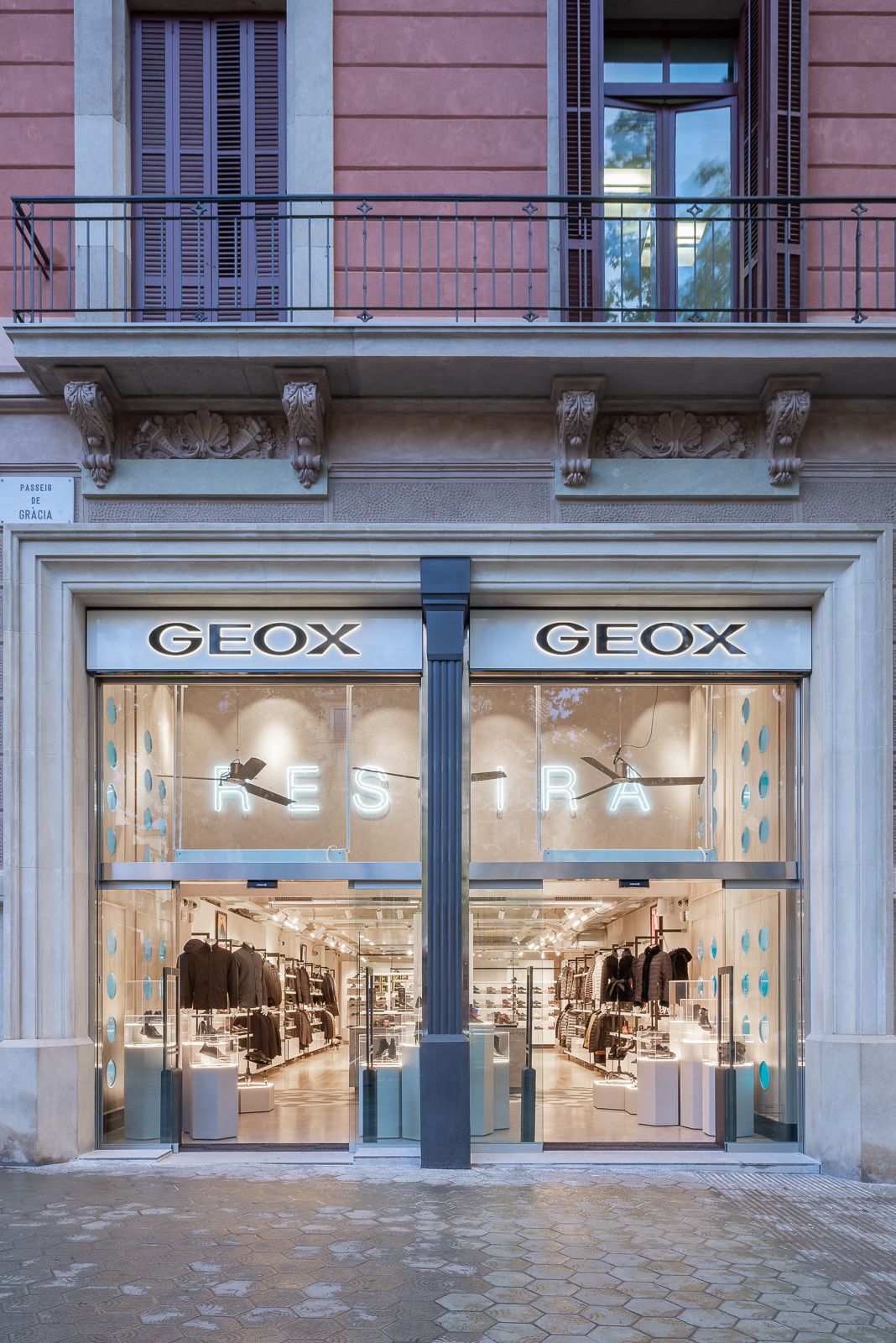 Vista frontal de una tienda Geox en Passeig de Gracia en Barcelona, con grandes ventanas de vidrio que muestran la mercancía interior y un balcón arriba.