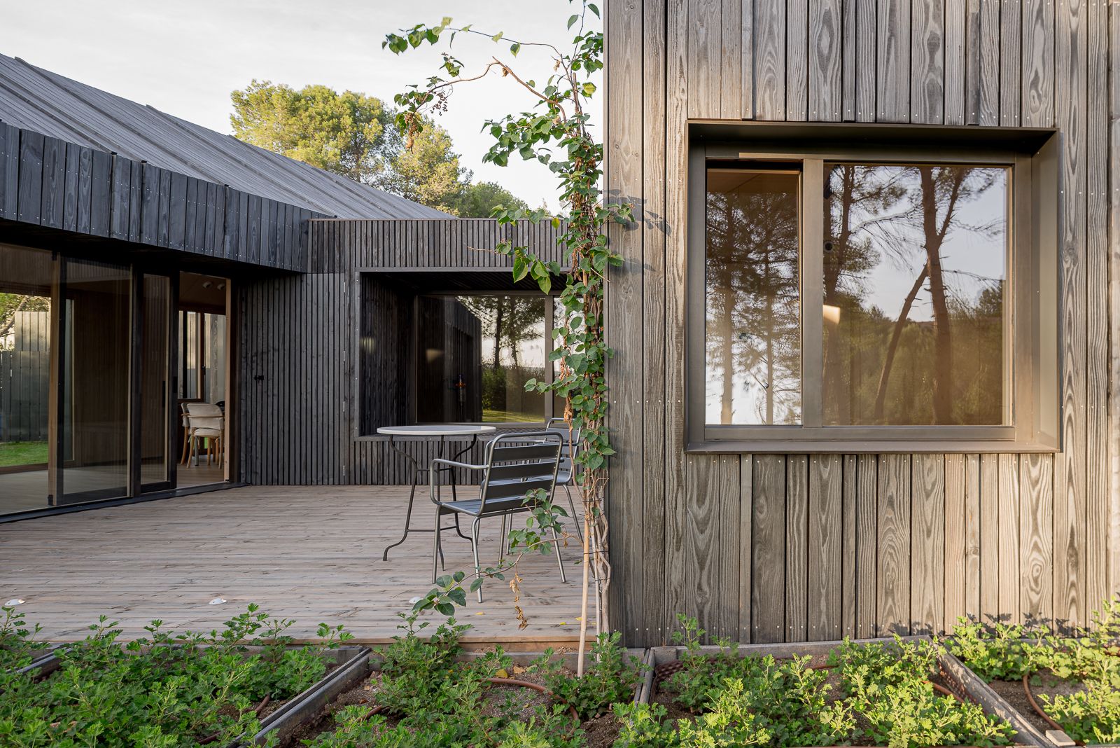Primer plano de una casa moderna con revestimiento de madera oscura, grandes ventanas y una terraza de madera con una pequeña mesa de mármol y sillas de metal.
