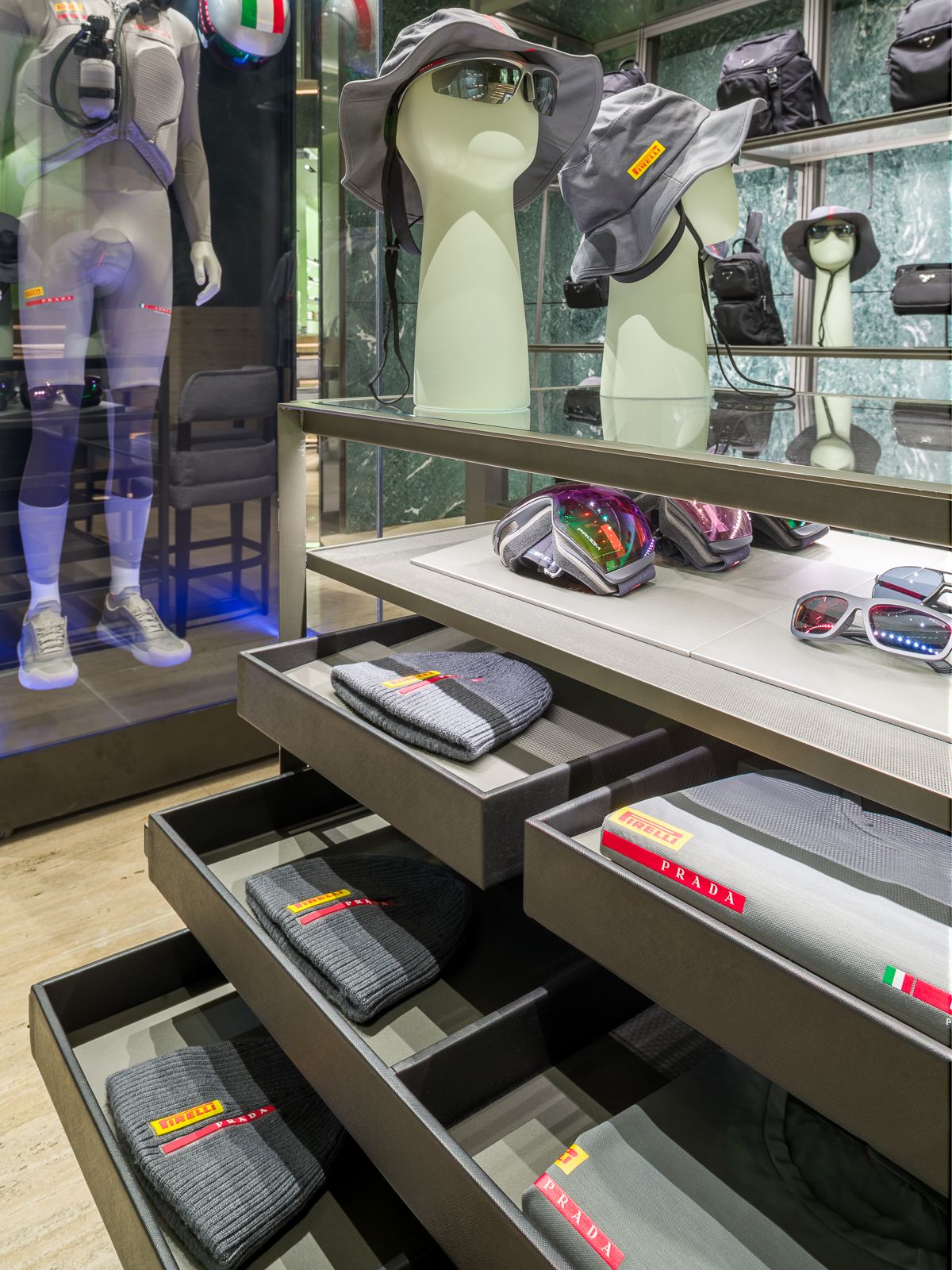 Lujosa boutique Prada, una mesa de vidrio con cajones metálicos medio abiertos mostrando prendas grises que exhiben las marcas Prada y Pirelli. Al fondo un maniquí en un atuendo de rendimiento está encerrado en una vitrina de vidrio.