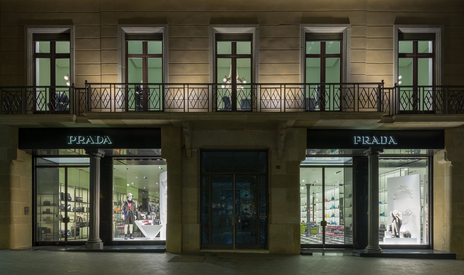 Exterior de una tienda Prada por la noche, mostrando escaparates brillantemente iluminados con artículos de lujo y maniquíes, enmarcados por una fachada de edificio clásico.