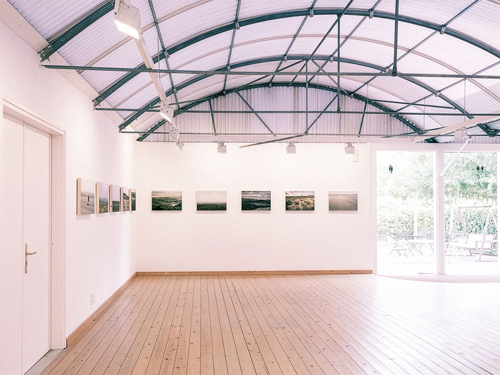 Gallery room with a curved ceiling and landscape photographs on the walls. Large door on the right open to a garden.