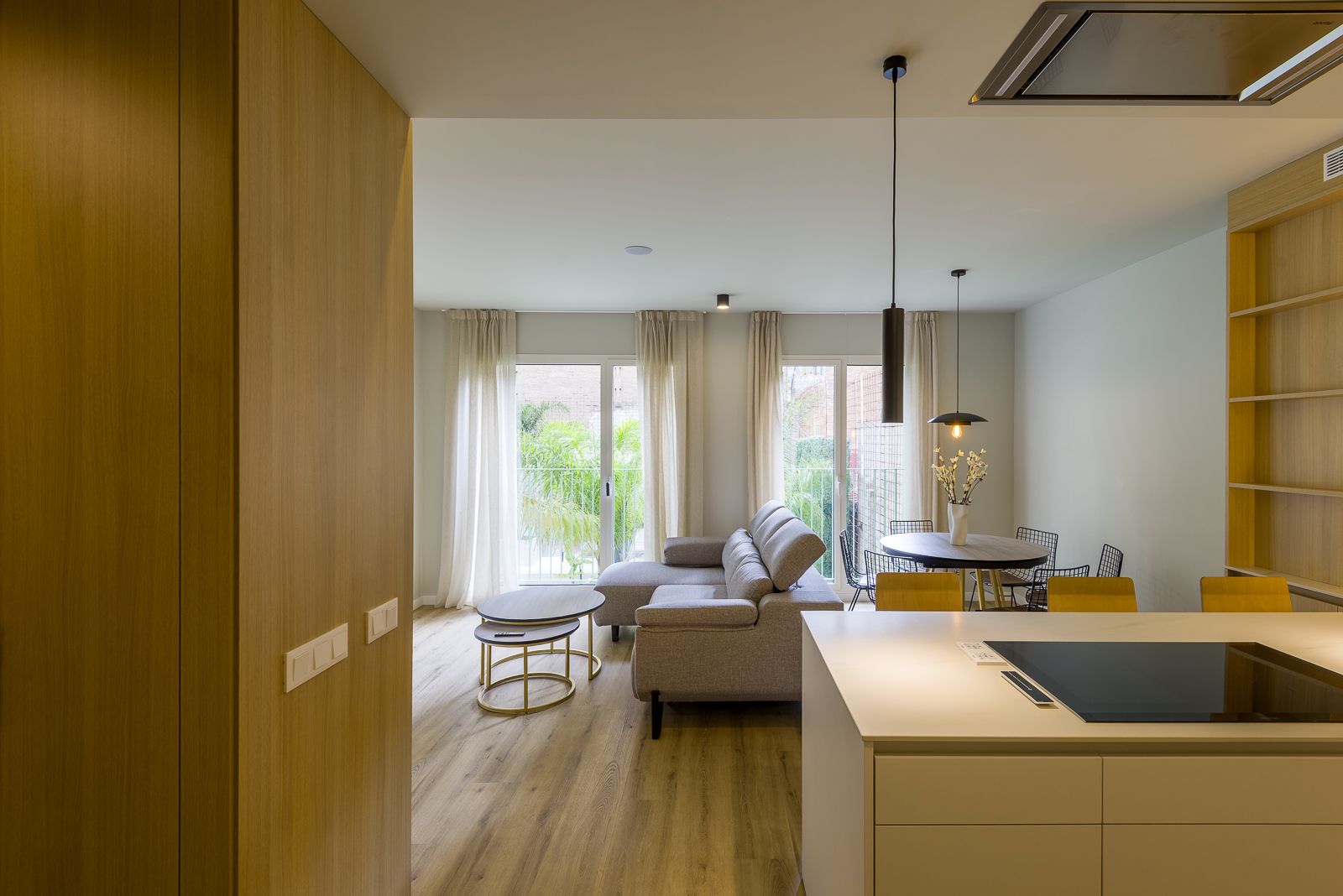 Bright, open-concept modern apartment seen from the kitchen, featuring light wood cabinetry, a beige sectional sofa, a small dining table with pendant lighting, and large balcony doors filtered by sheer white curtains.